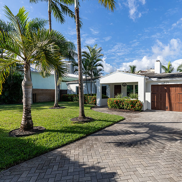 Singlestory white house with a wooden garage door palm trees and a brick driveway on a sunny day Single-story white house with a wooden garage door, palm trees, and a brick driveway on a sunny day.