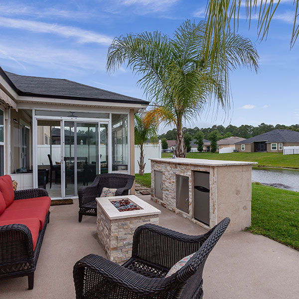 Outdoor patio with wicker seating around a rectangular fire pit and builtin grill beside a glass sunroom and palm trees with a pond in the distance Outdoor patio with wicker seating around a rectangular fire pit and built‑in grill, beside a glass sunroom and palm trees, with a pond in the distance.