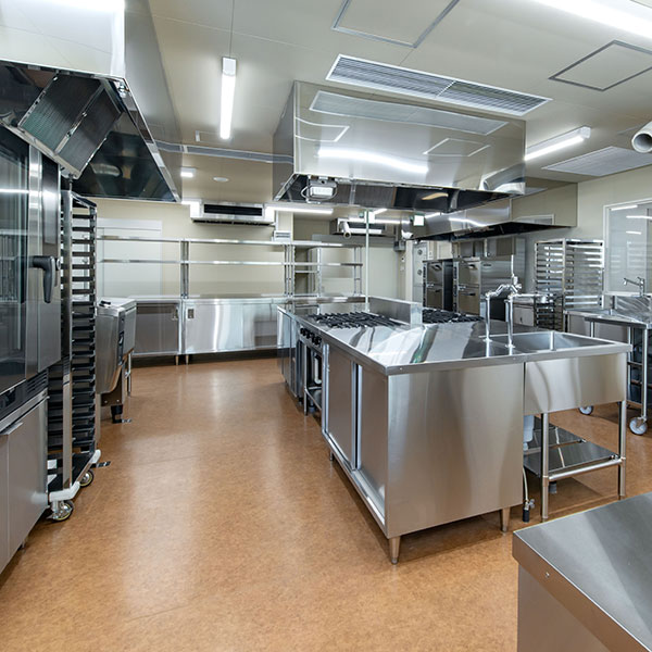 Commercial stainless steel kitchen with central prep island gas range and overhead vent hoods all illuminated by bright ceiling lights Commercial stainless steel kitchen with central prep island, gas range, and overhead vent hoods, all illuminated by bright ceiling lights.